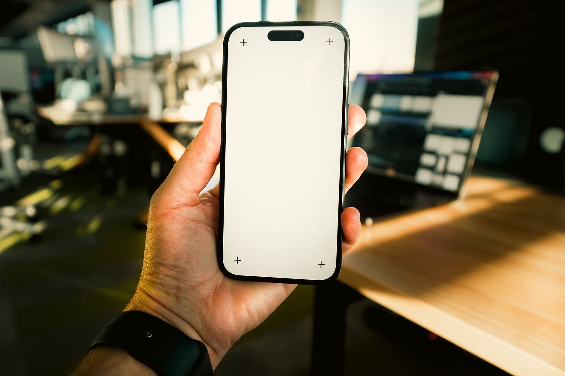 Close-up of hand holding smartphone with blank screen in a sunny office setting.