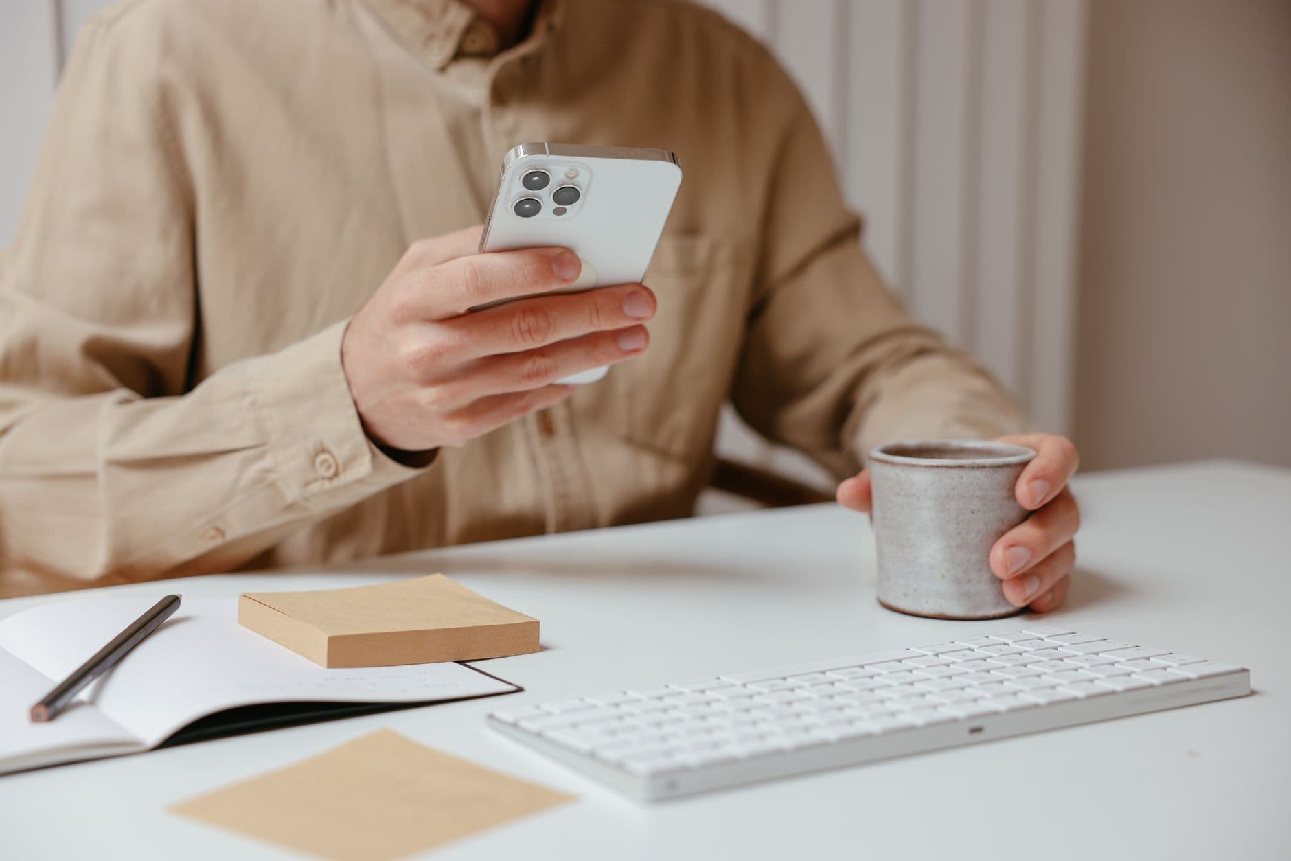 A man holding a smartphone and cup while sitting at a desk with keyboard and notepad.