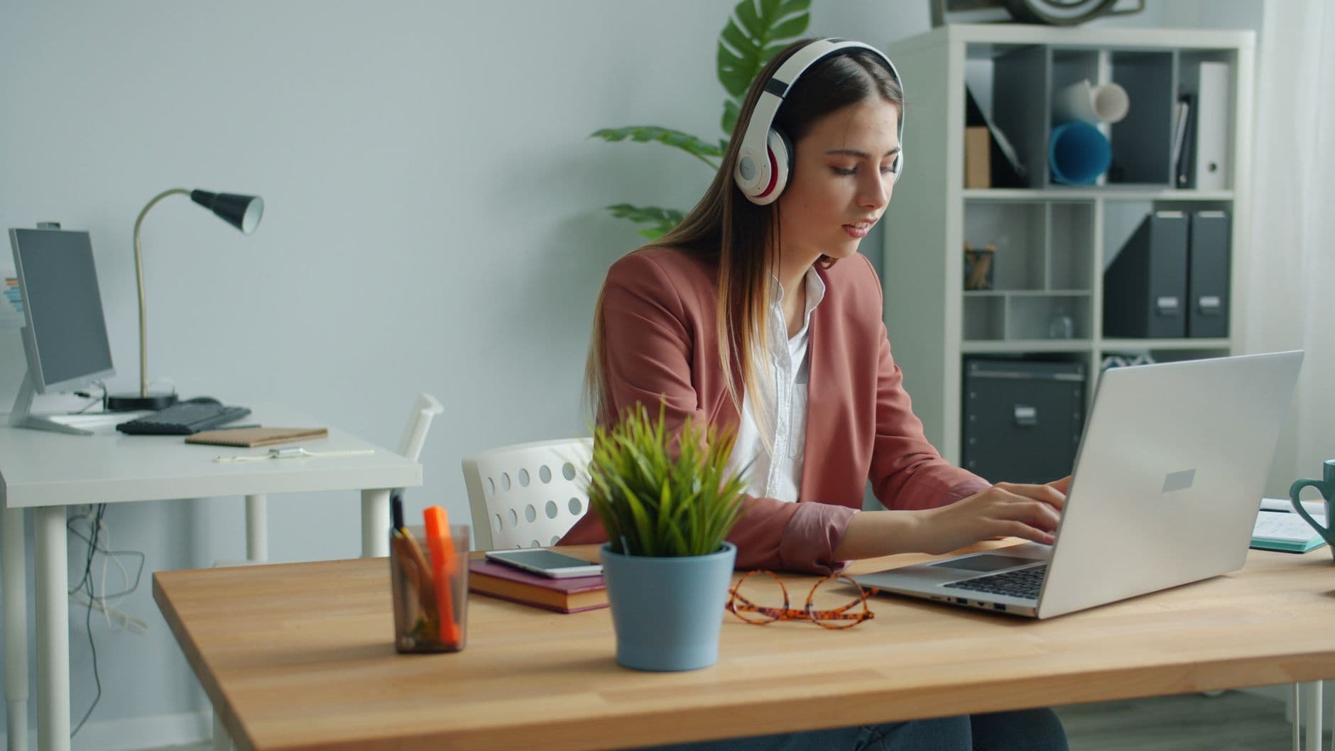 Focused young woman wearing headphones while working remotely on her laptop from a home office.
