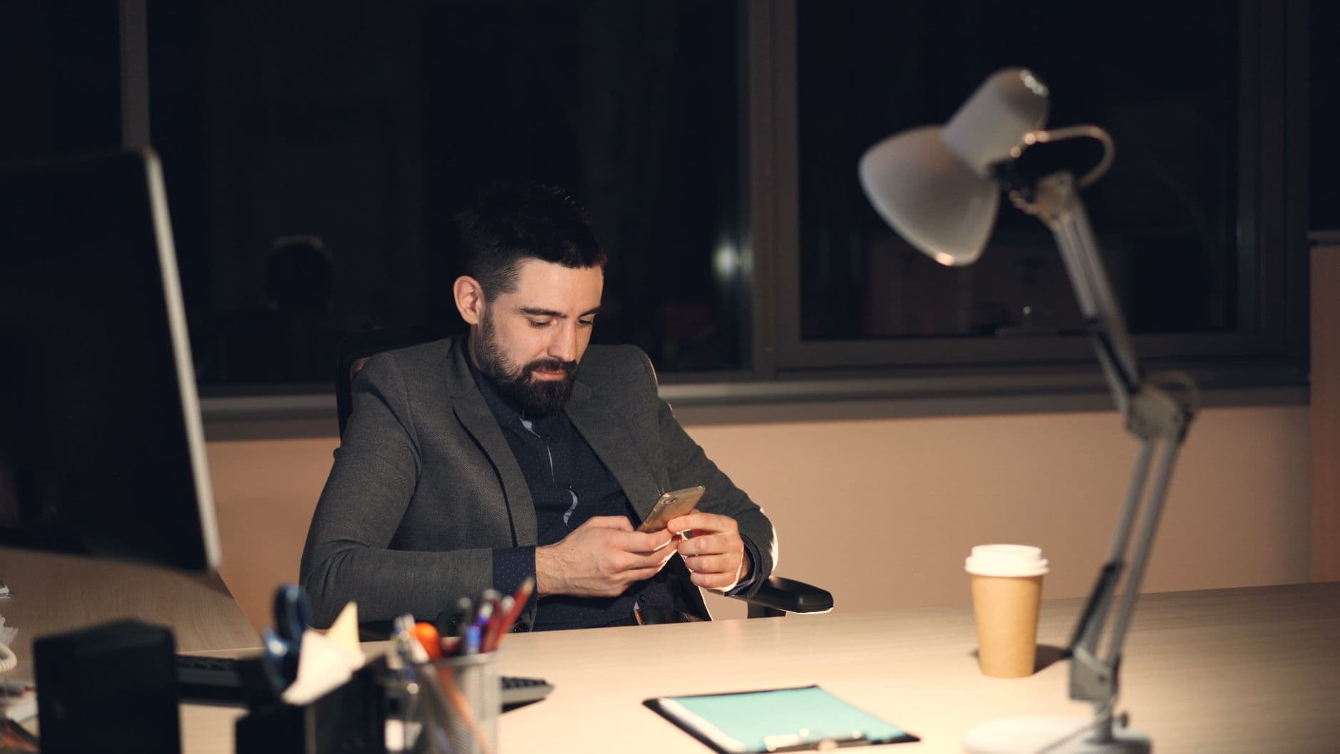 A businessman in a suit using a smartphone at his office desk at night.