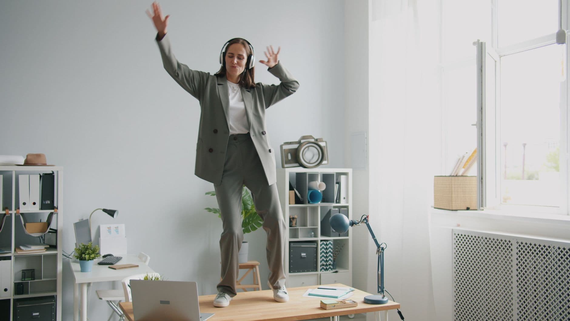 Businesswoman in suit and headphones dancing energetically on office desk.