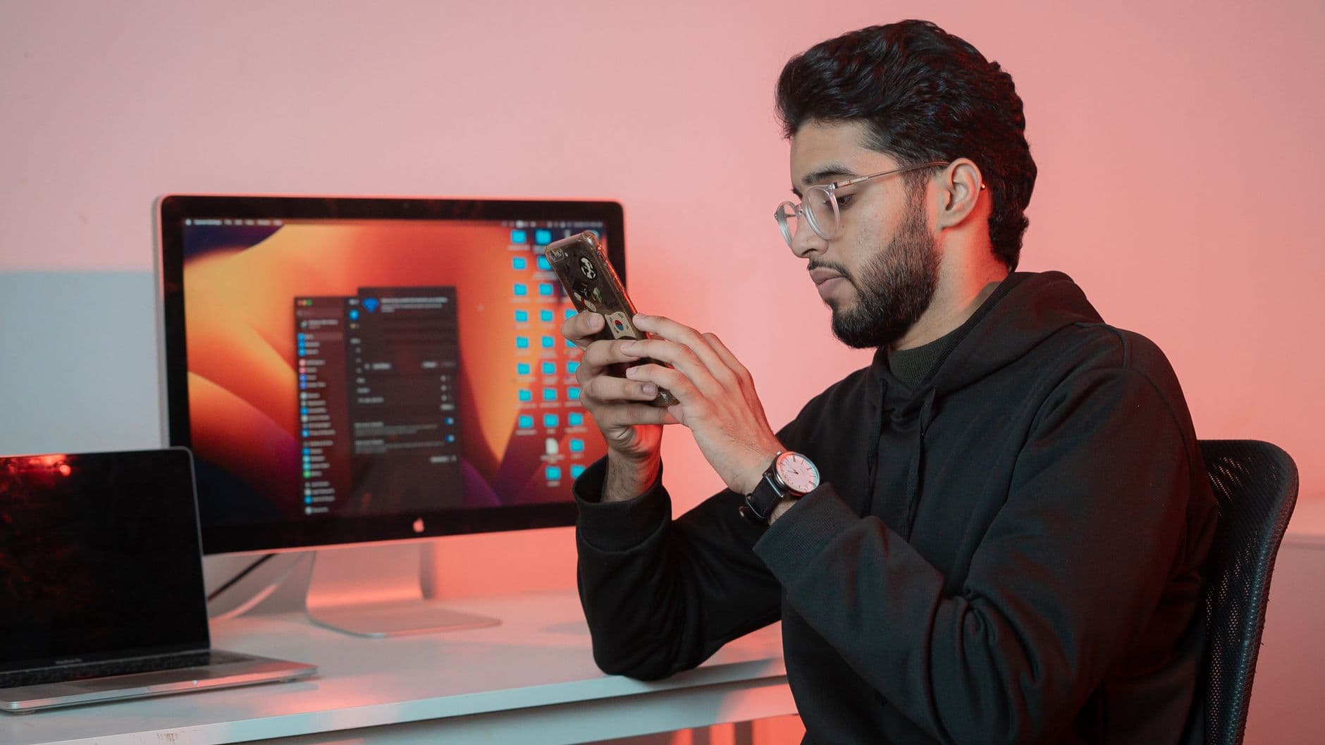 Adult male using smartphone while working at desk with computer in modern office setting.