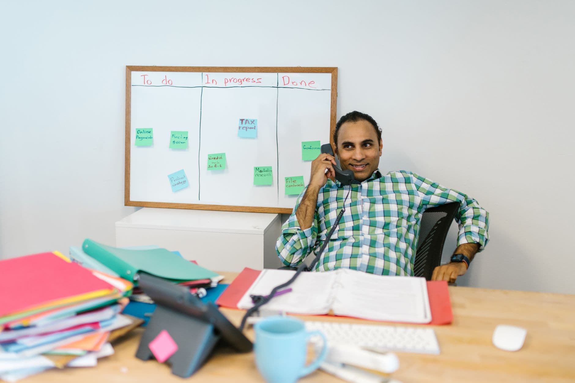 An adult male in a modern office setting, working at a desk with a task board in the background.