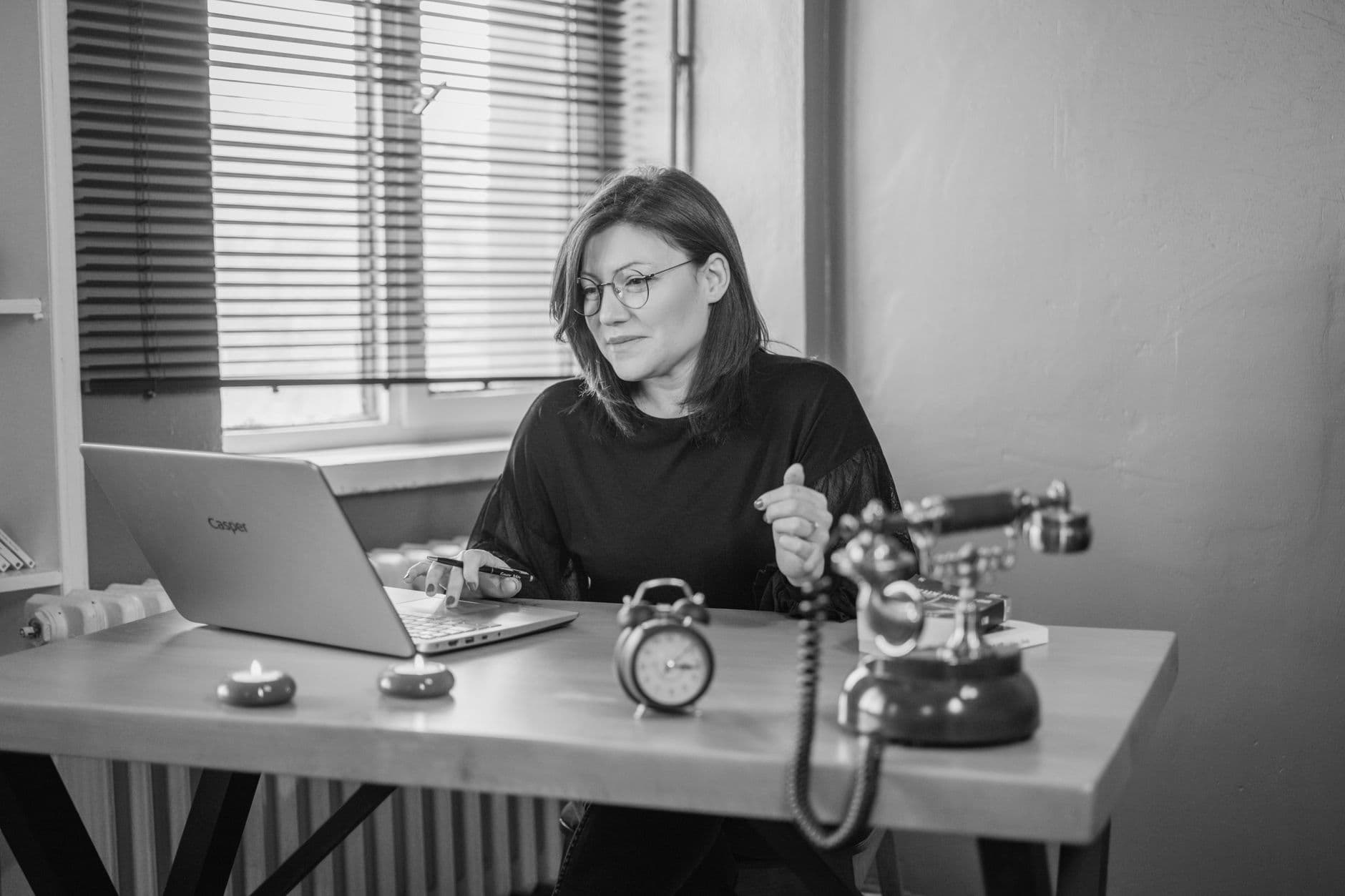 Black and white photo of a woman working at a desk with a laptop and vintage telephone.